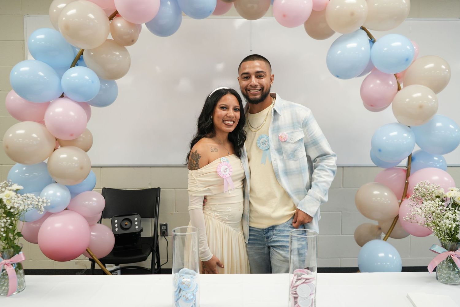 Couple smiling together at a gender reveal party under a pink, blue, and gold balloon arch.