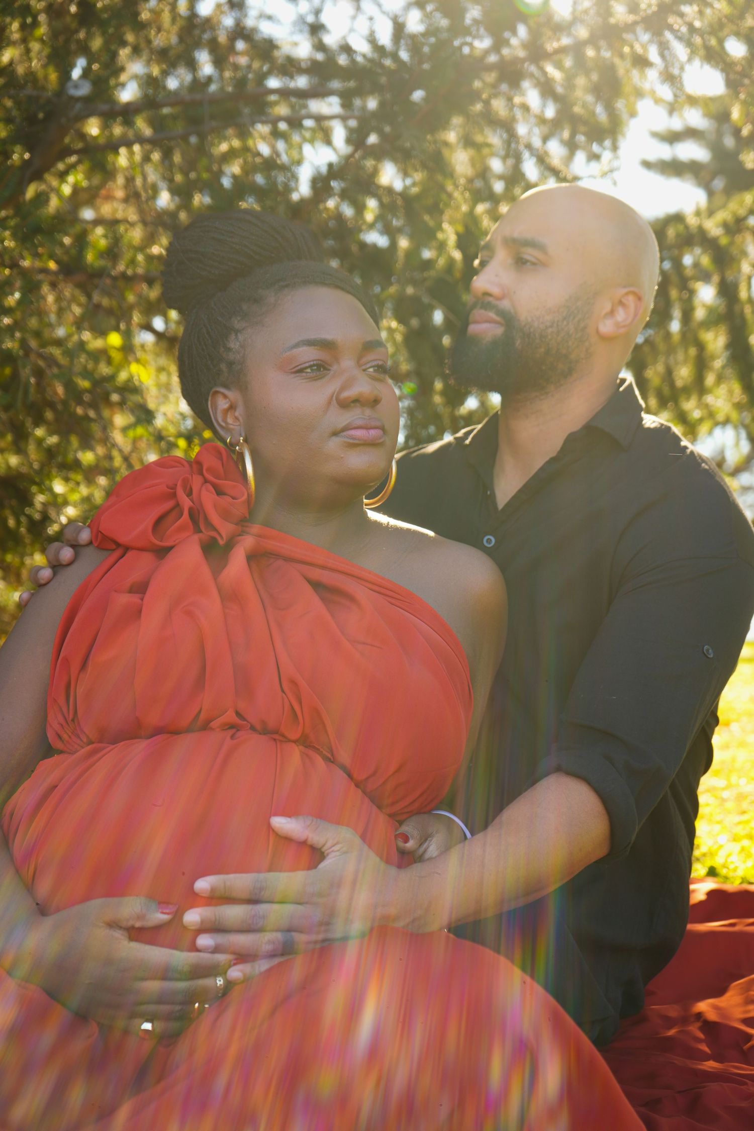 Pregnant woman in red dress with partner outdoors in golden sunlight, couple maternity photo session in park.