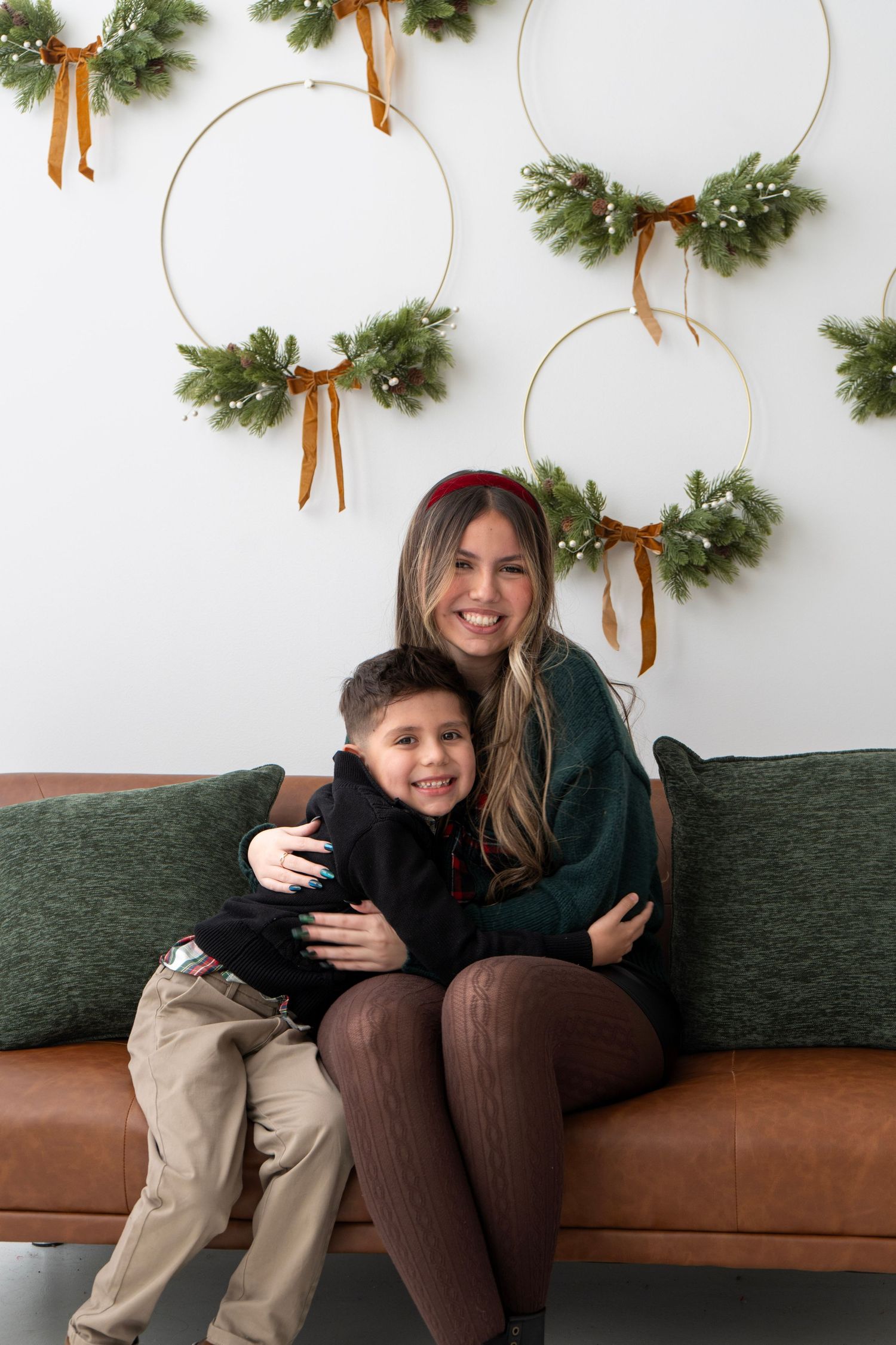 A smiling woman hugs a young boy on a leather sofa with festive Christmas wreath decorations on the wall behind them.