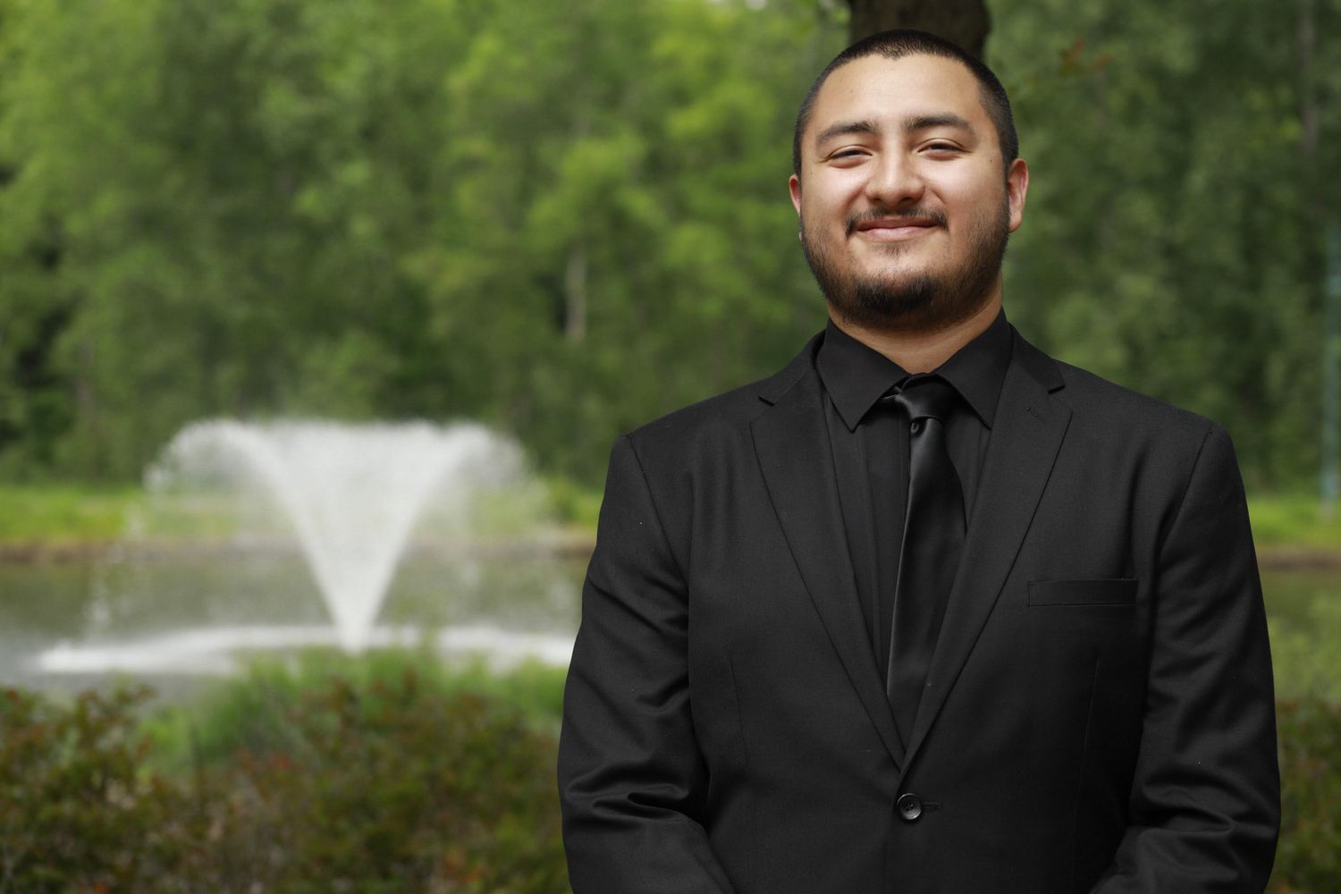 Man in black suit and tie smiling outdoors near a decorative fountain surrounded by lush green trees.