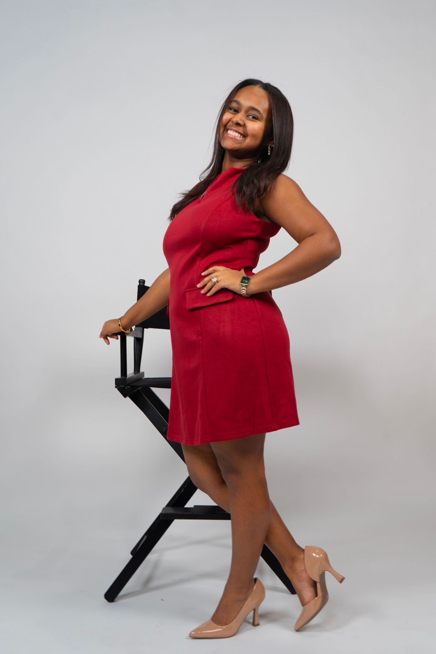 Woman in red dress and nude heels posing confidently beside a black director's chair against gray background.