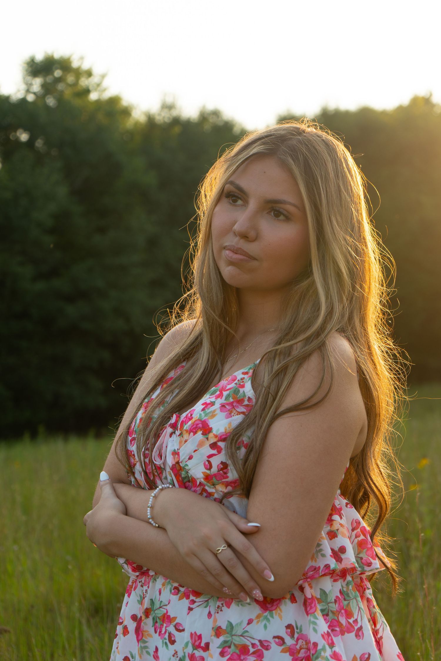 Young woman in floral dress with arms crossed standing in sunlit green field at golden hour.