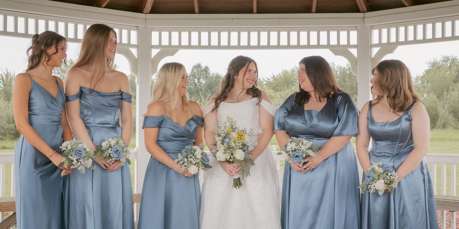 Bridesmaids in dusty blue dresses holding floral bouquets pose together under a white gazebo.