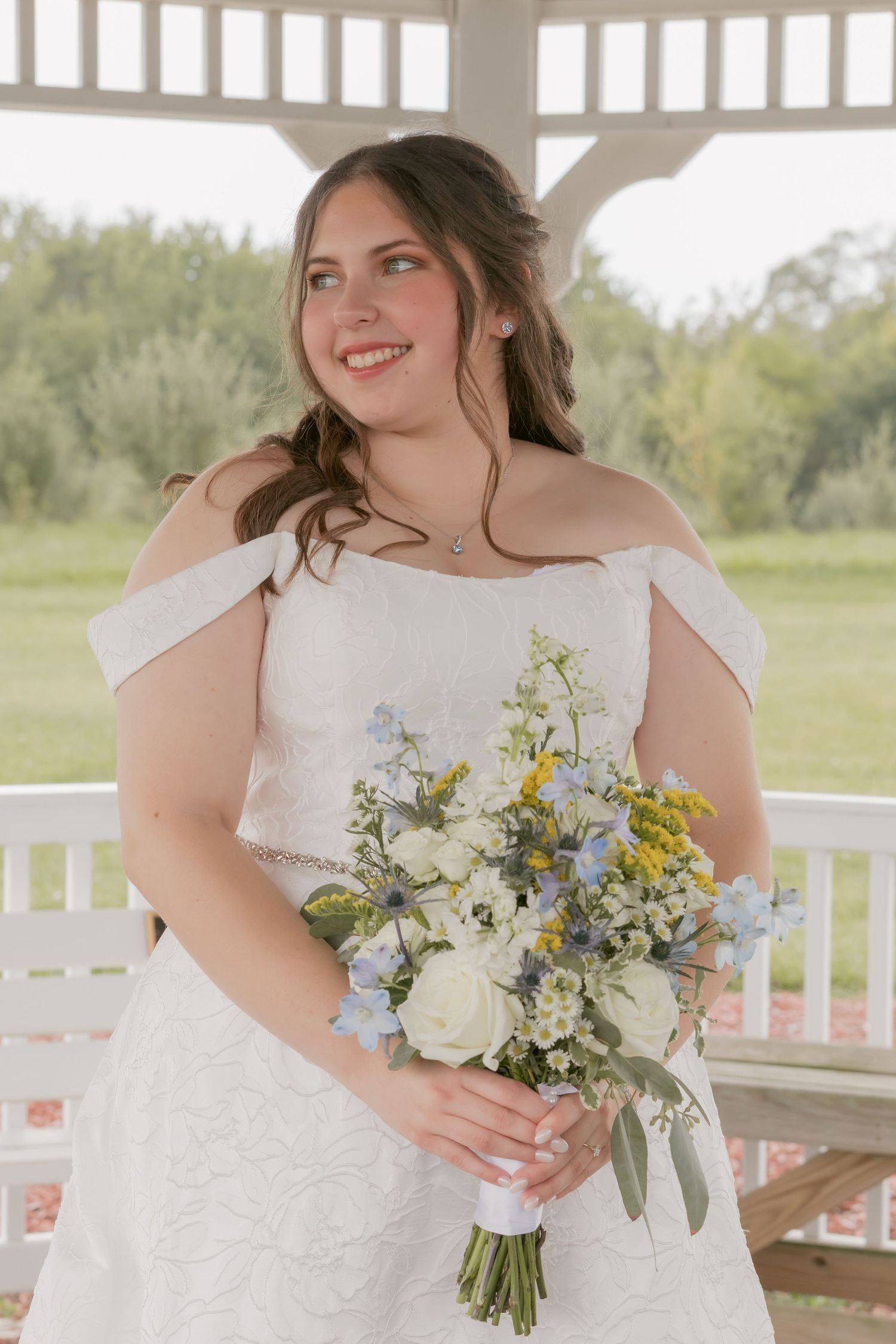 Bride in off-shoulder wedding dress holds a colorful bouquet while posing in a gazebo.
