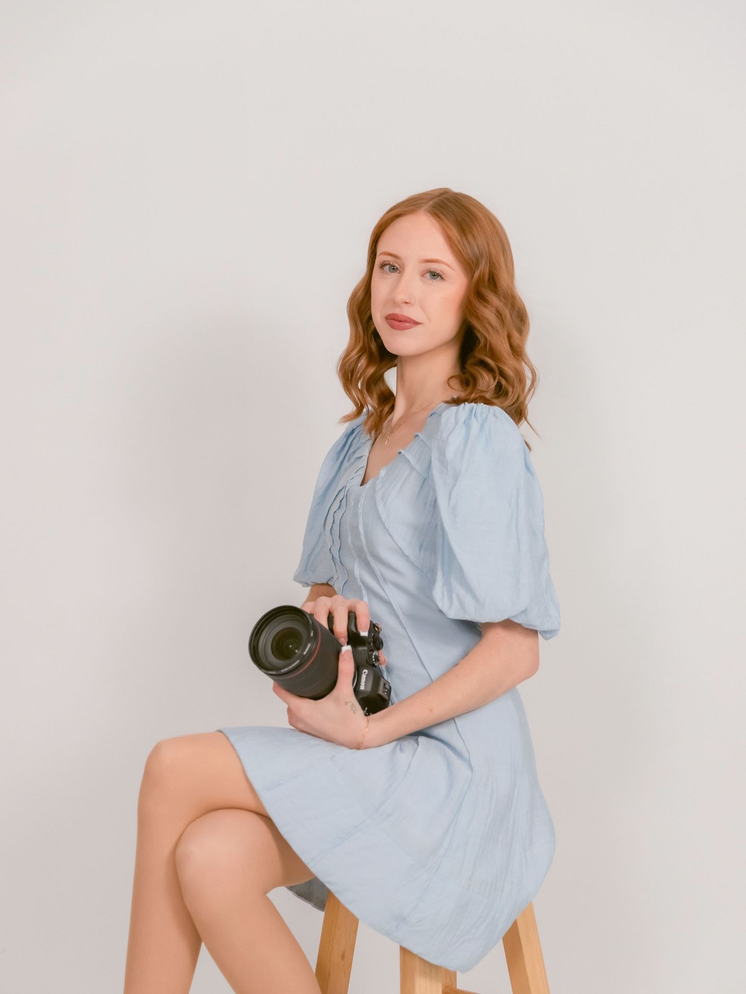 Person in powder blue dress poses with camera on wooden stool against white background.