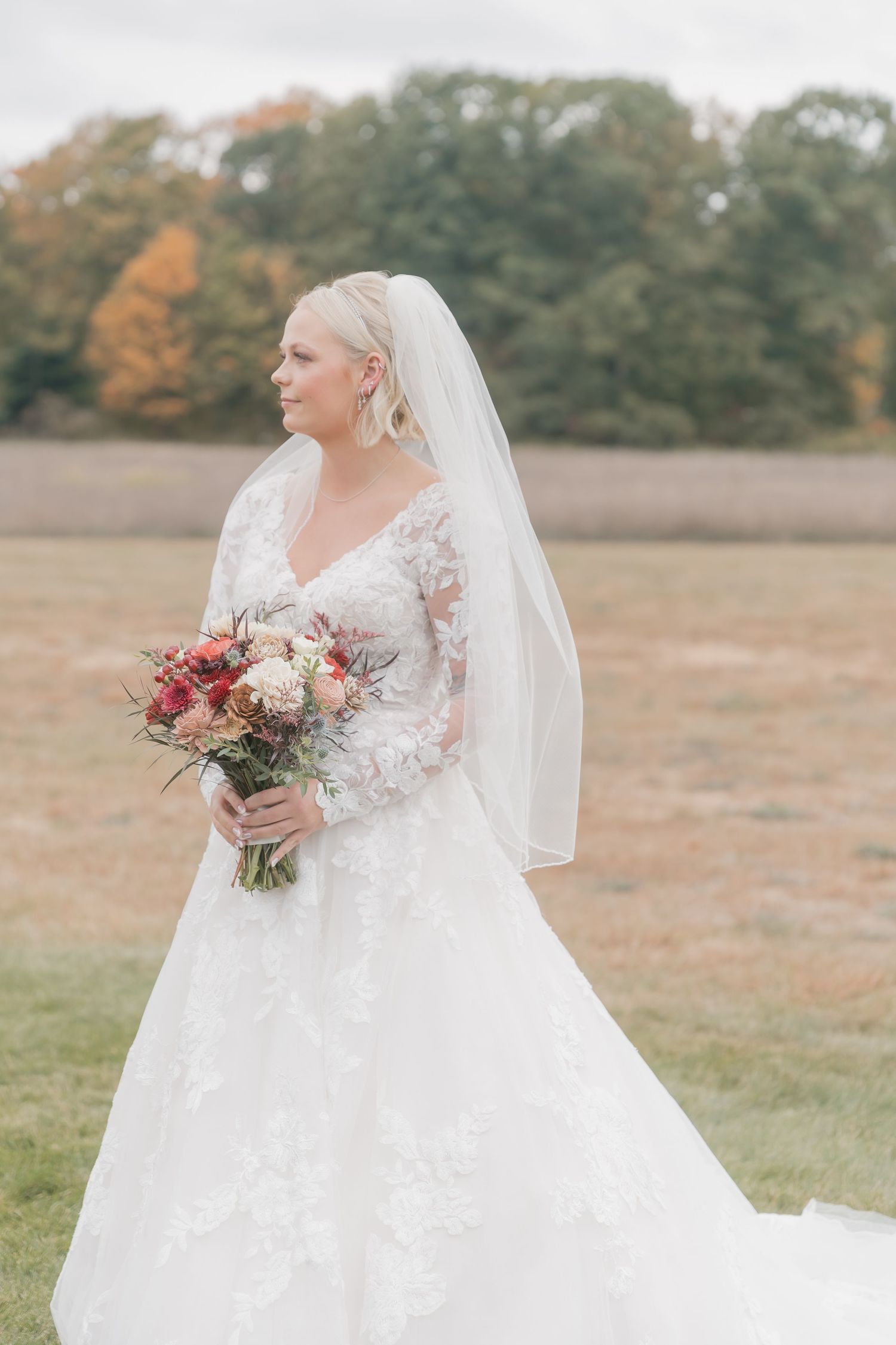Bride in detailed lace gown holds autumn-colored bouquet outdoors.