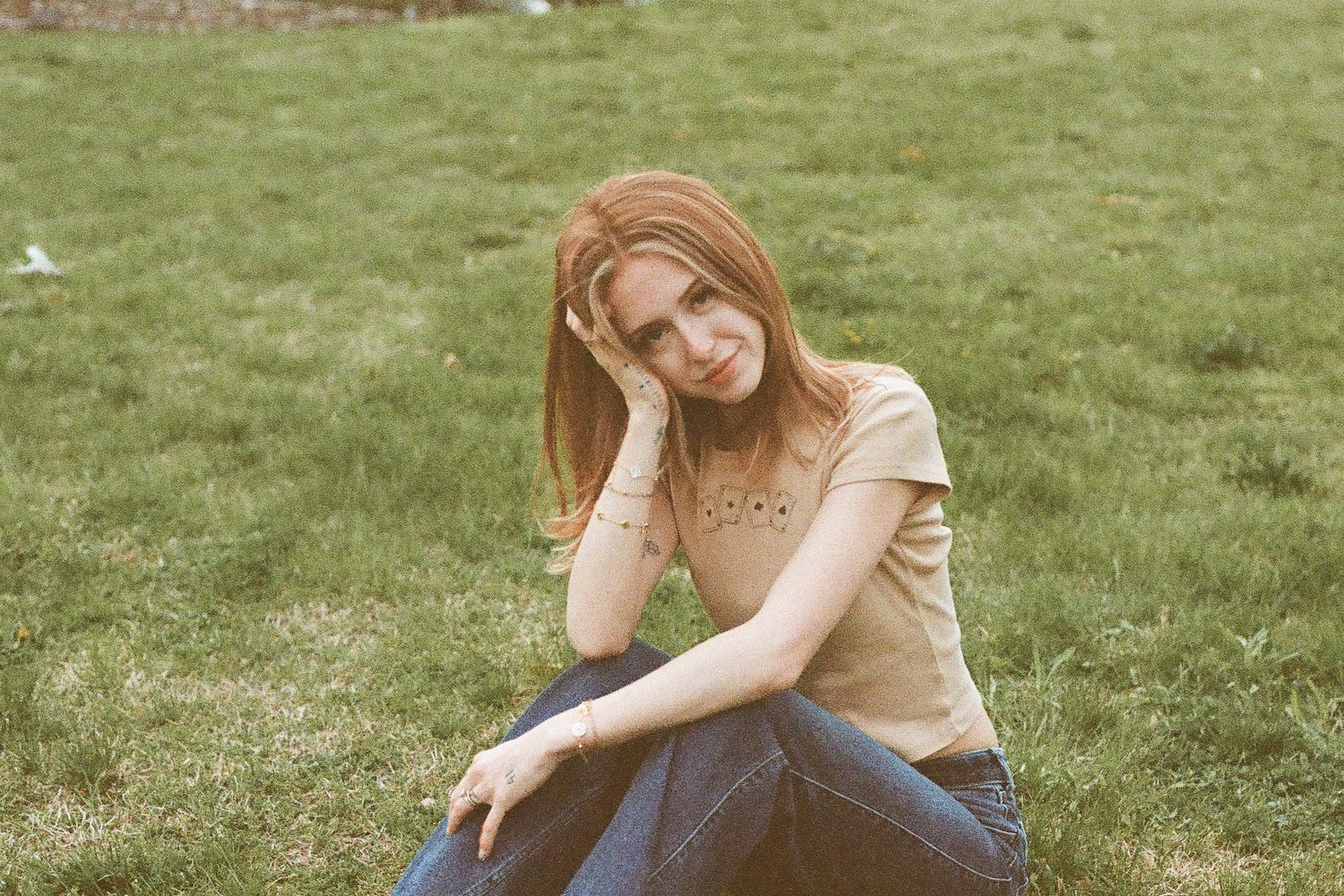 Young woman with red hair sitting on green grass, wearing a tan top and jeans, resting her head on her hand.