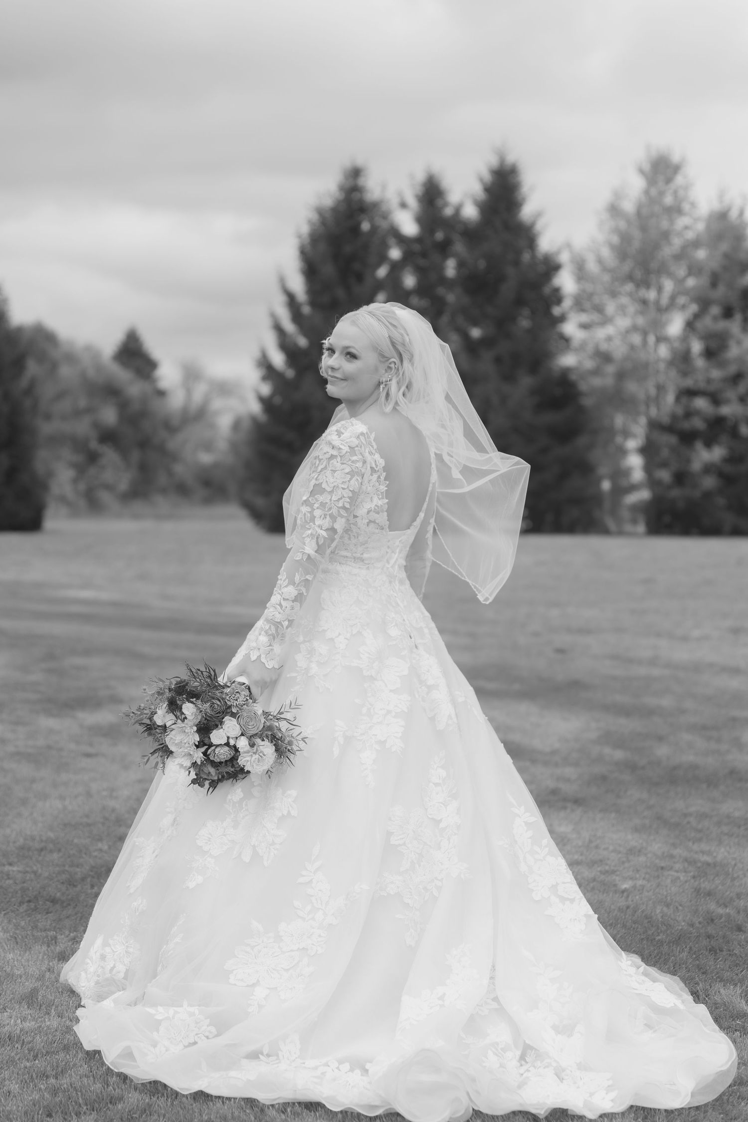 Black and white portrait of a bride in a lace gown with flowing veil in an outdoor setting.