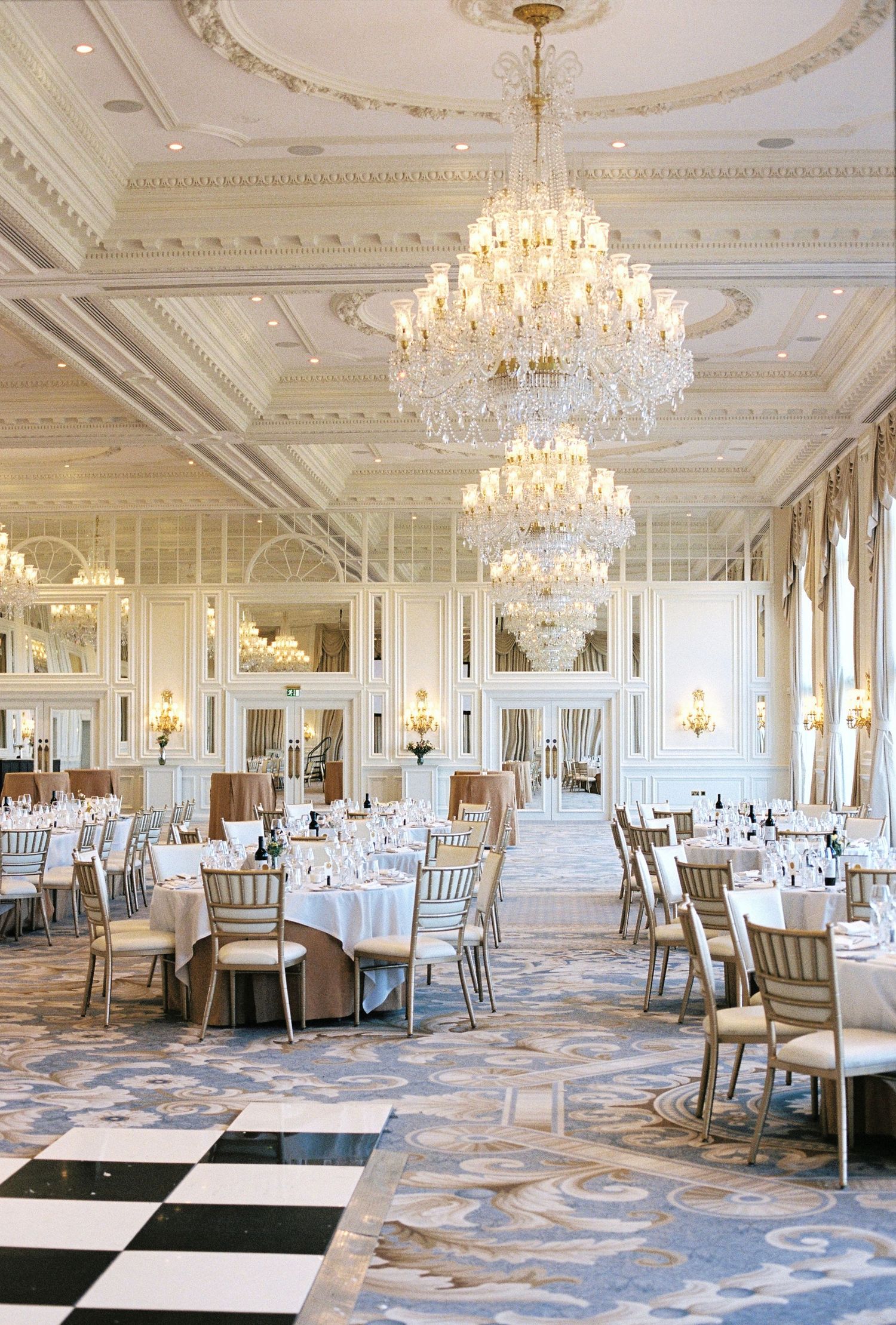 Elegant ballroom with crystal chandeliers, white decor, round tables, gold chairs, and a black-and-white checkered dance floor.
