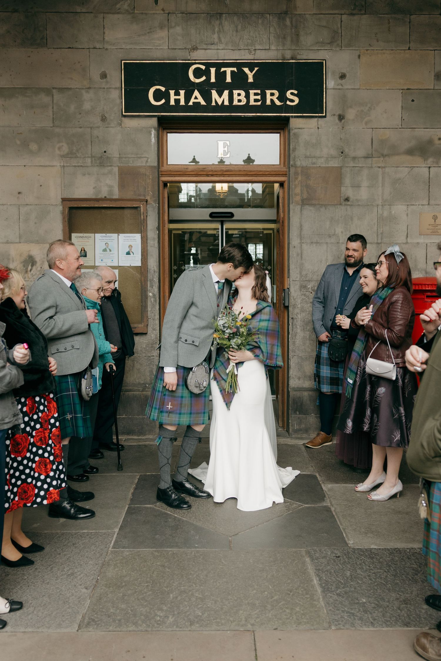 Newlyweds kiss outside Edinburgh City Chambers, groom in tartan kilt, bride in white gown, guests cheering around them.