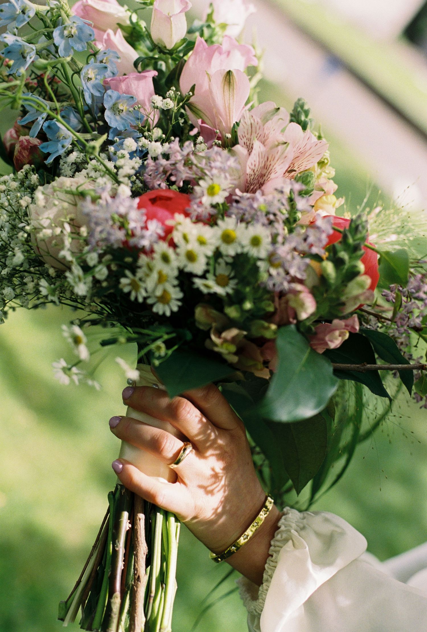 Close-up of bride holding lush wedding bouquet with pink, white, and red flowers, wearing gold bracelet.