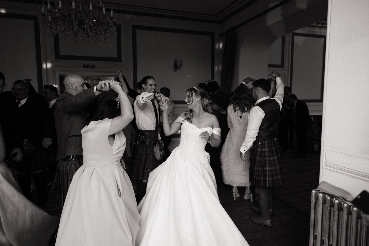 Bride and guests dancing joyfully at a Scottish wedding reception in a grand ballroom, black and white.