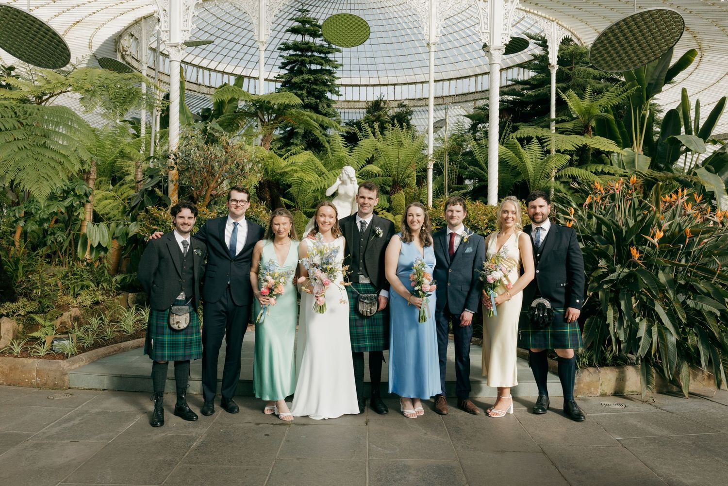Wedding party posing in a stunning glass botanical garden with lush tropical plants, wearing kilts, blue dresses, and bridal gown.