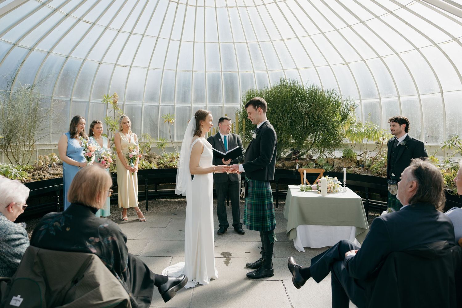 Wedding ceremony in a glass greenhouse, bride in white gown and groom in tartan kilt exchanging vows before guests.