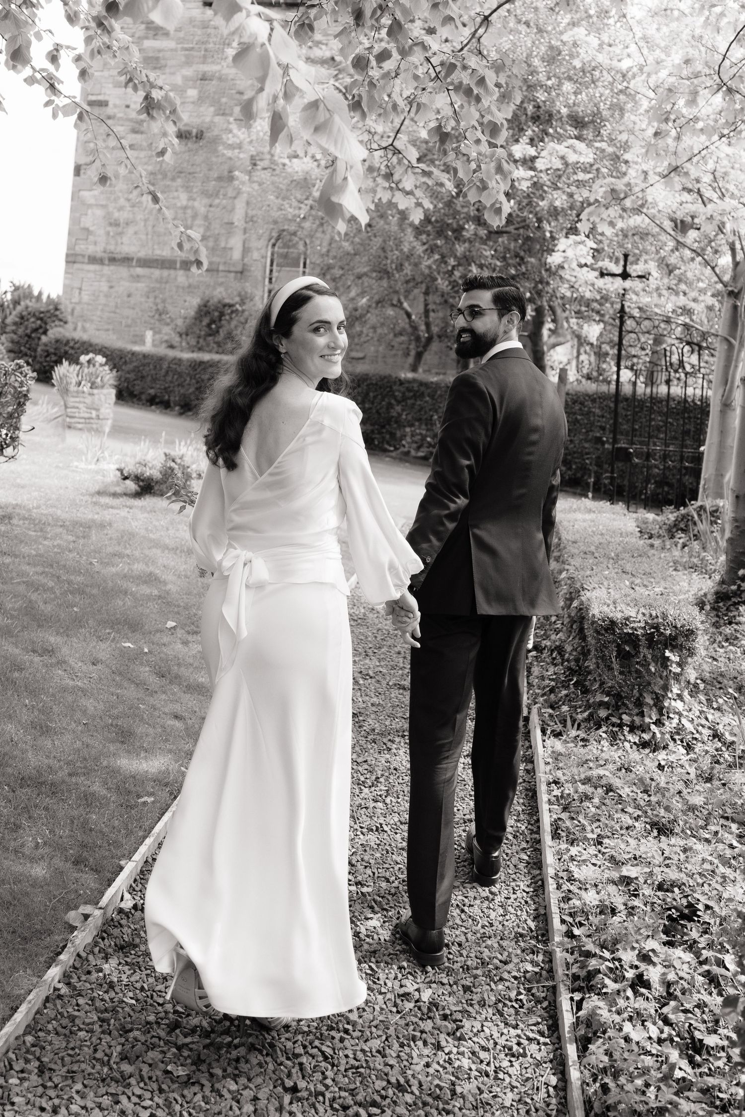 Black and white photo of a bride and groom walking hand in hand through a garden, bride glancing back.