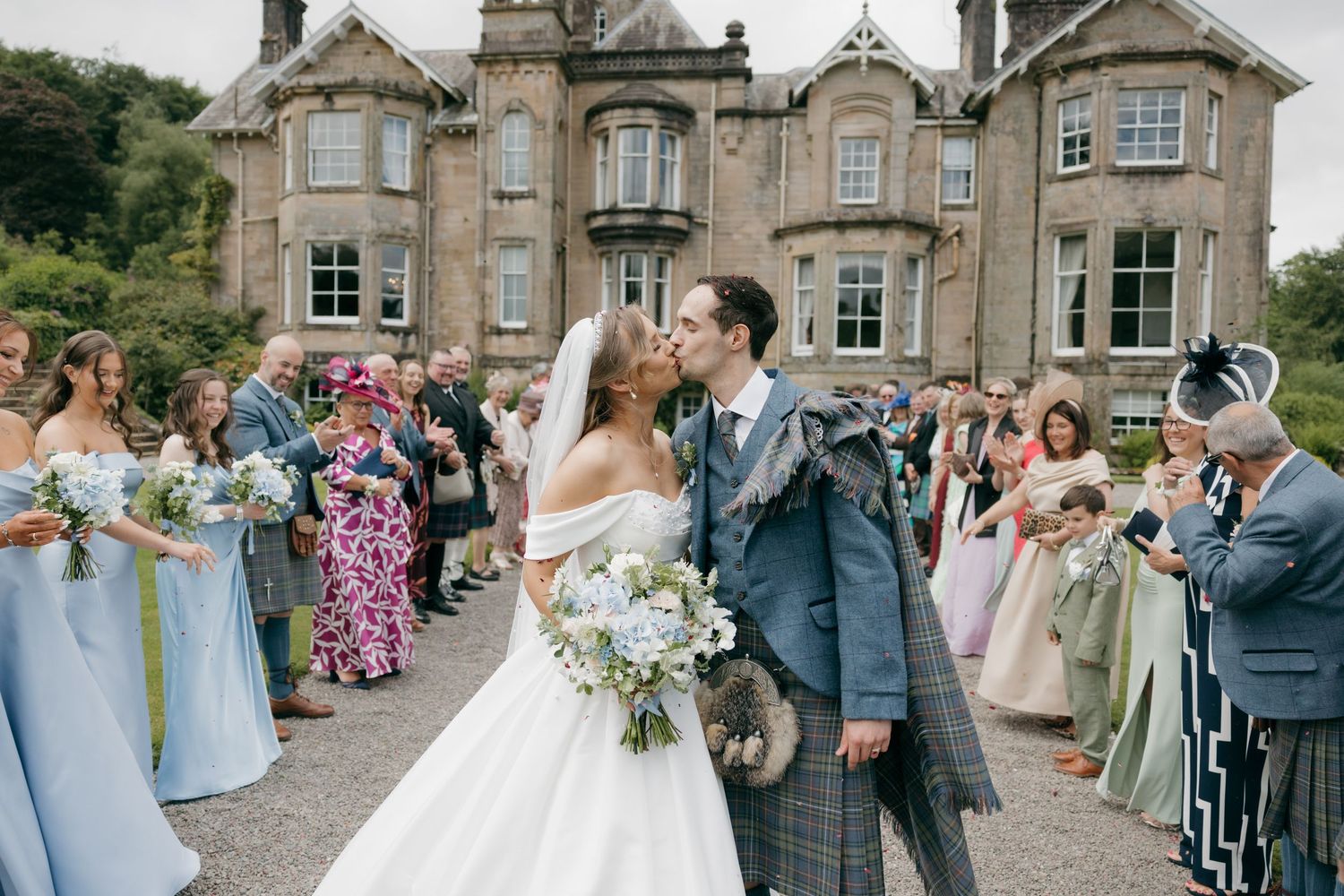 Bride and groom share a kiss outside a Scottish castle wedding venue surrounded by guests and bridesmaids in blue dresses.