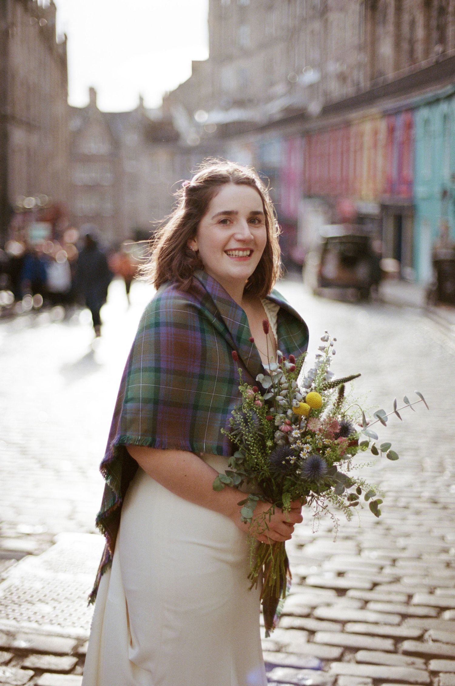 Smiling bride in white dress and tartan shawl holding wildflower bouquet on cobblestone street in Edinburgh.
