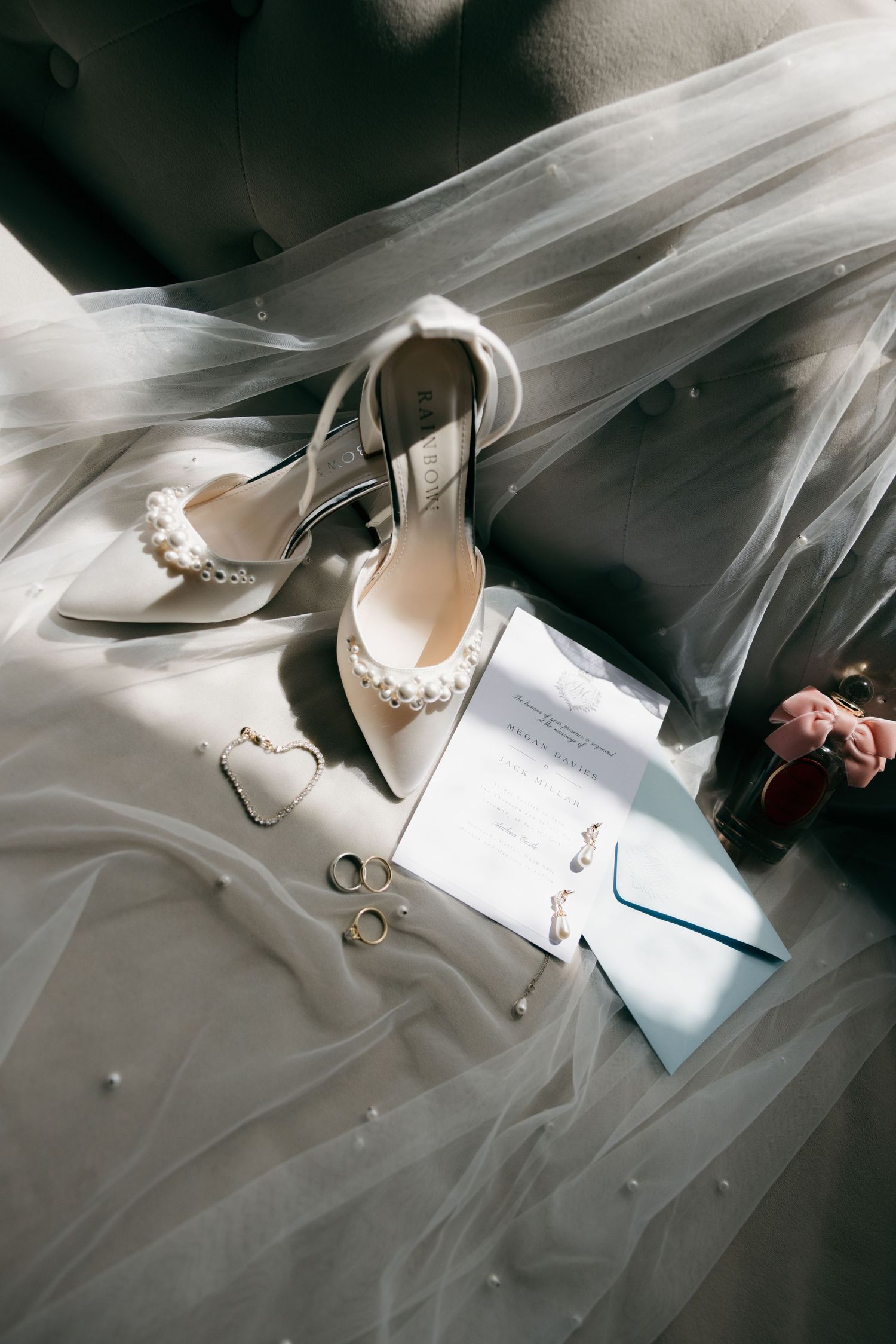 Wedding flat lay with pearl heels, veil, rings, earrings, and invitation in natural light.