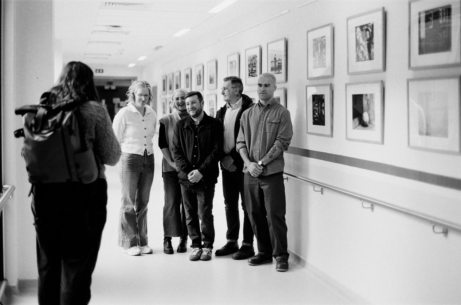 Black and white photo of people posing in a hallway gallery while being filmed by a cameraman.