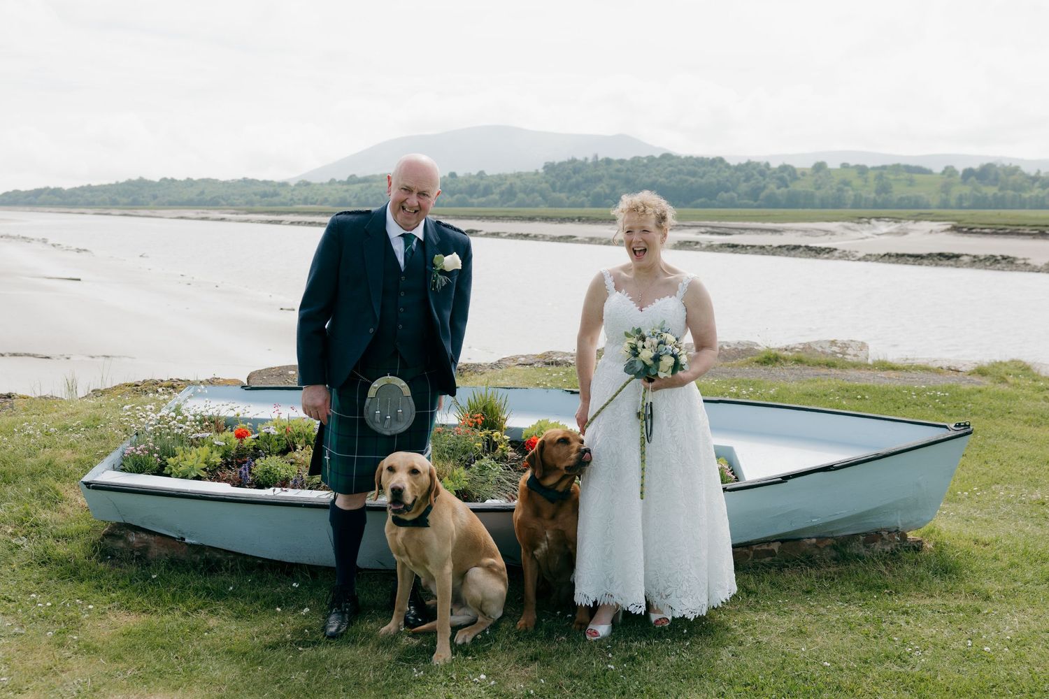 Bride and groom in Scottish kilt pose with two golden dogs beside a decorative boat with loch and mountains behind.