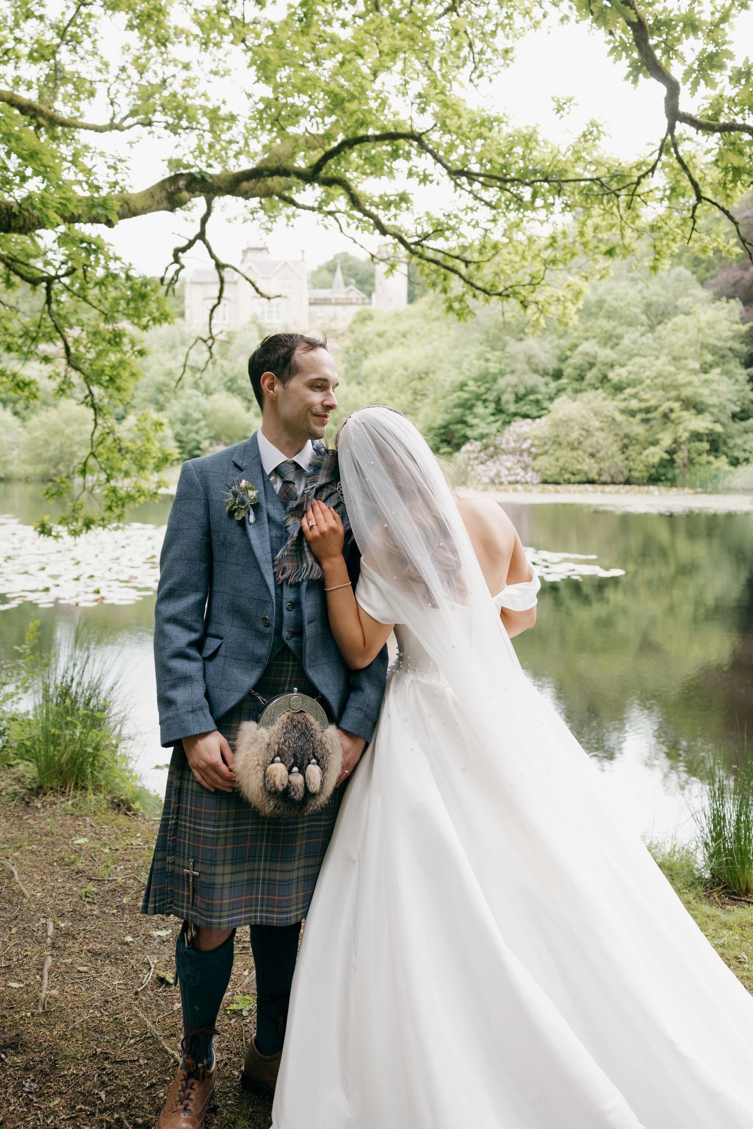 Groom in blue Scottish kilt suit smiles at bride by tranquil lake beneath lush green trees.