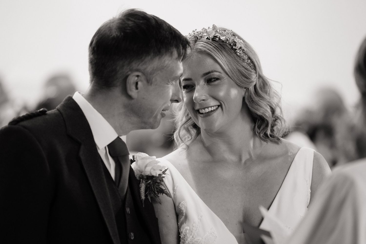 Black and white wedding photo of a smiling bride in floral crown gazing at her groom in a suit.