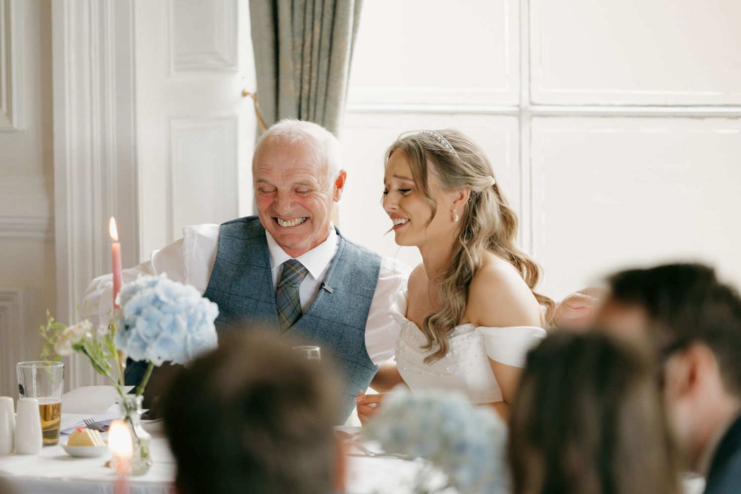 Bride in white off-shoulder gown laughs with older gentleman in blue suit at elegant wedding reception table.