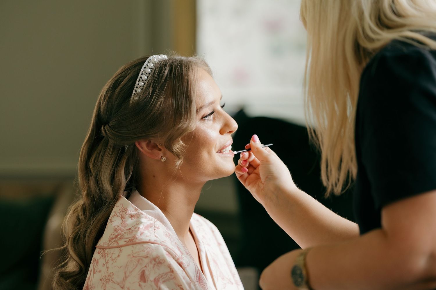 A makeup artist applying lipstick to a smiling bride wearing a floral robe and hair accessory.