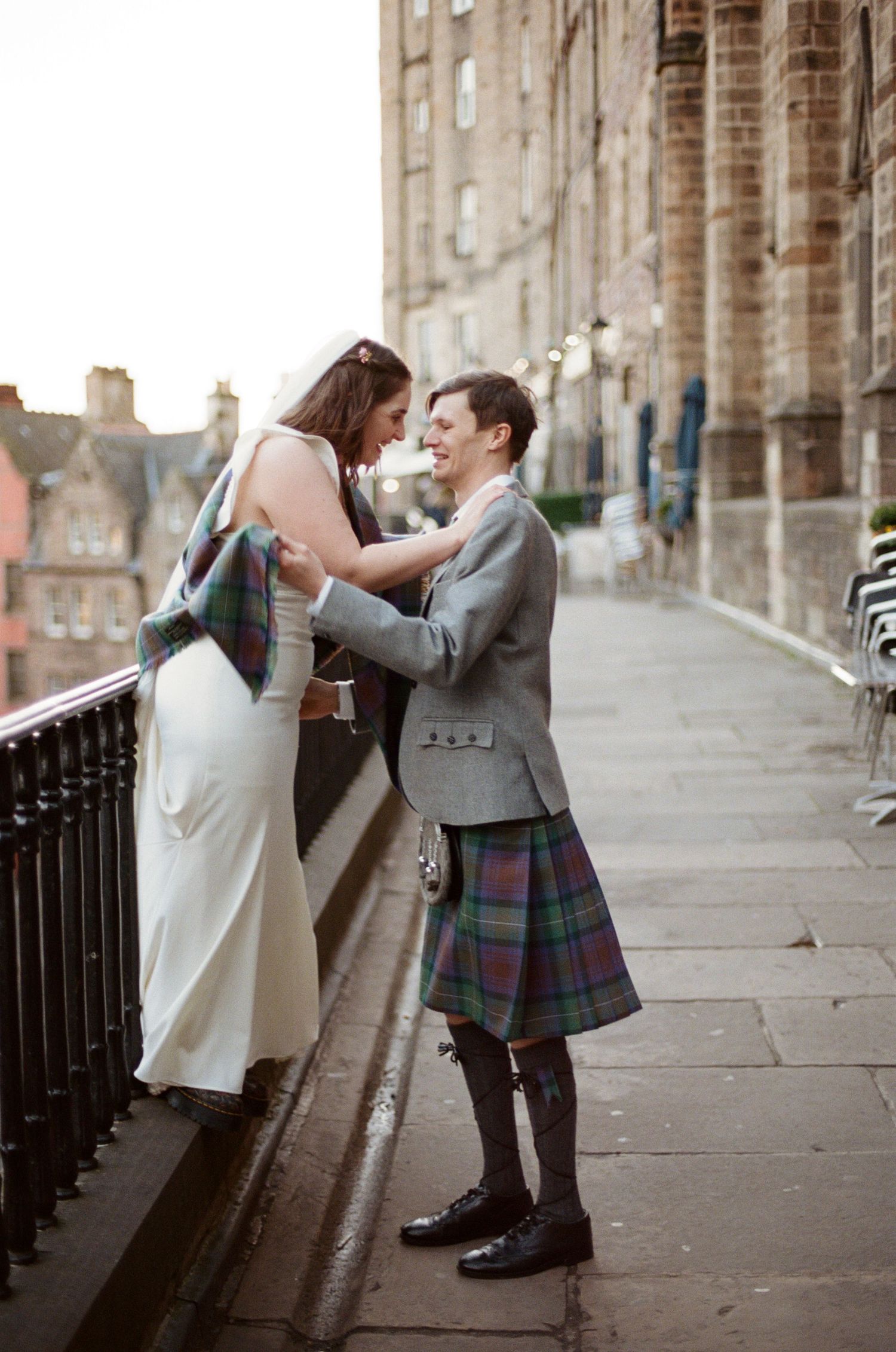Bride and groom in Scottish tartan kilt share romantic moment on Edinburgh cobblestone street at golden hour.