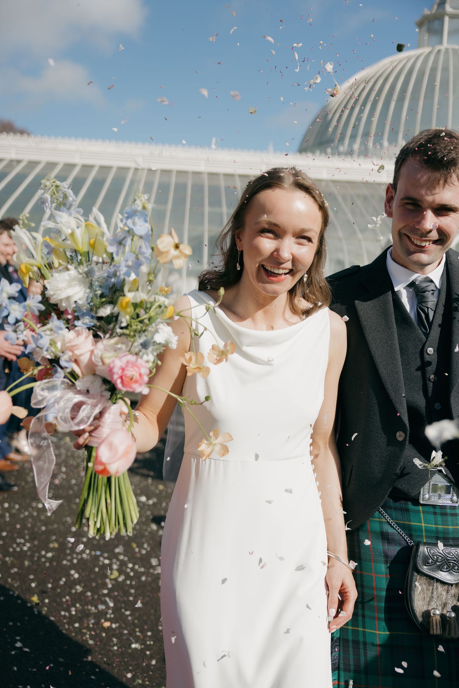 Joyful bride in white gown holding pastel bouquet with groom in tartan kilt, confetti shower outside glass greenhouse.