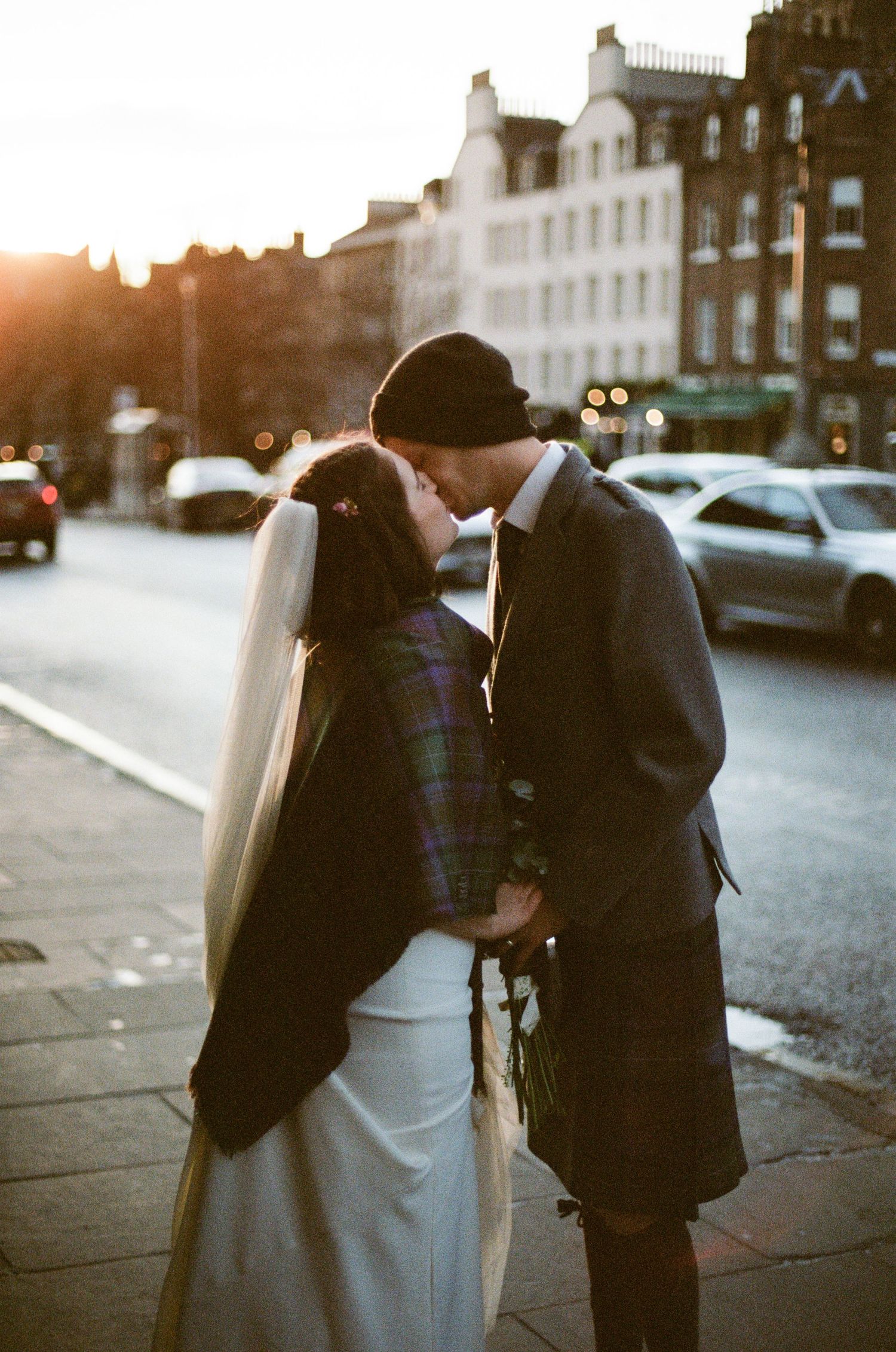A bride and groom share a romantic kiss on a cobblestone street at golden hour, with the groom wearing a traditional Scottish kilt.