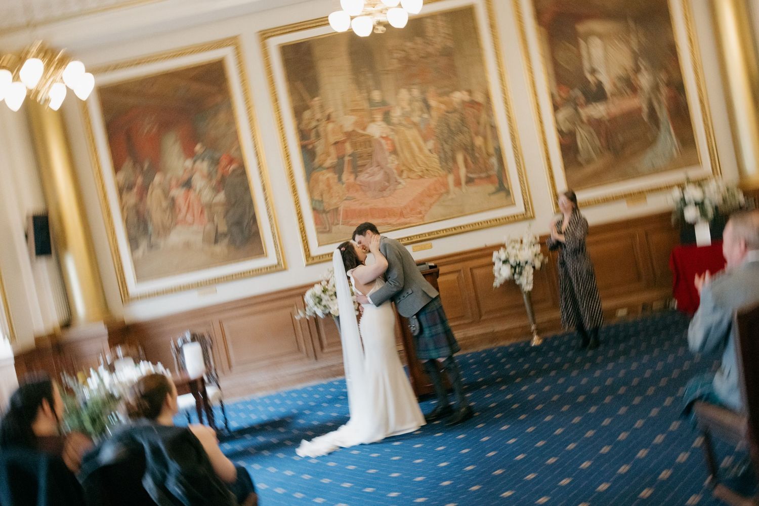 Bride and groom share first kiss at elegant wedding ceremony in ornate blue-carpeted hall with classical paintings.