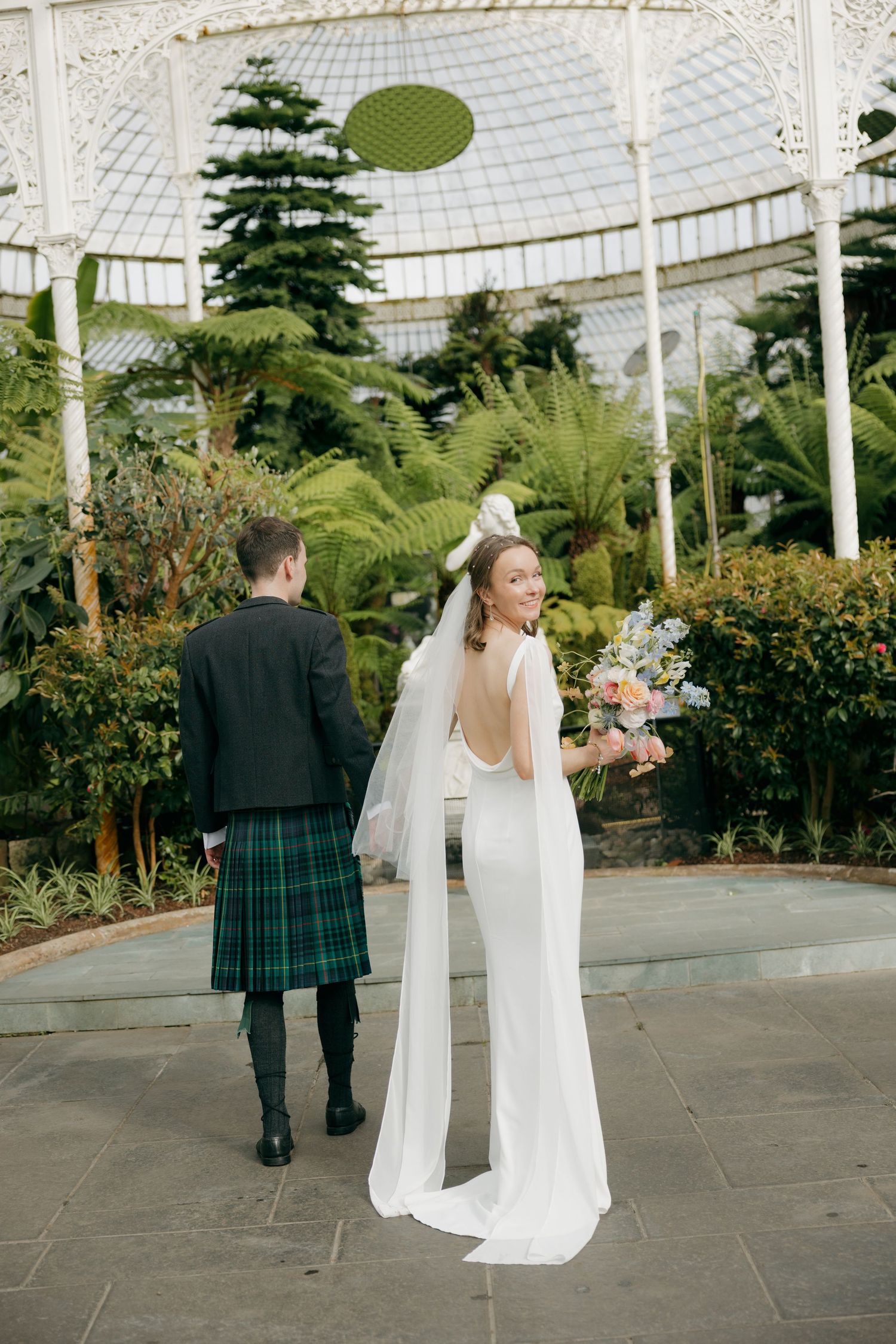 Bride and groom in kilt standing in botanical garden greenhouse on wedding day.