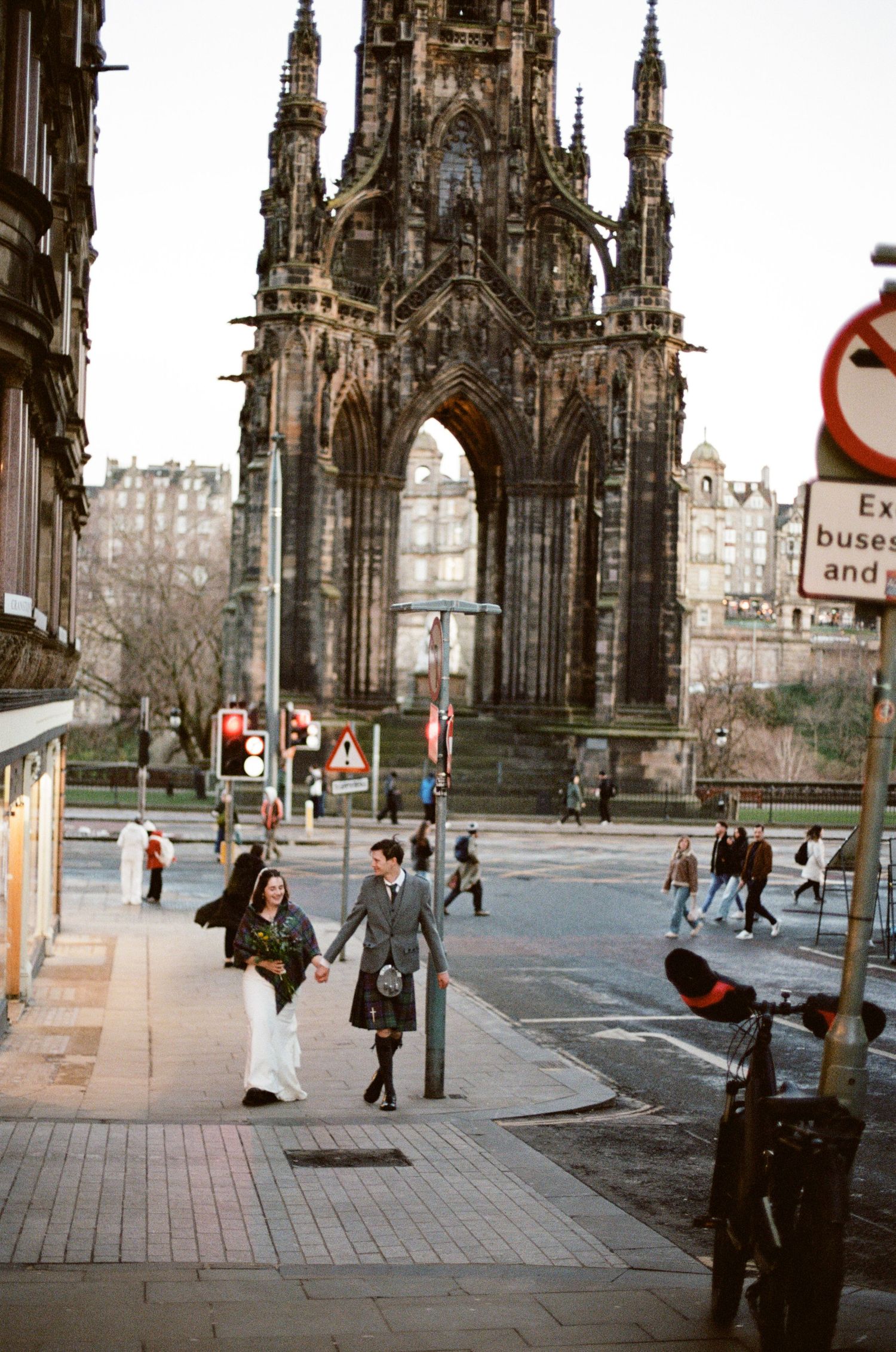 Wedding couple in Scottish attire walking near the Scott Monument in Edinburgh, groom in tartan kilt, bride in white dress.