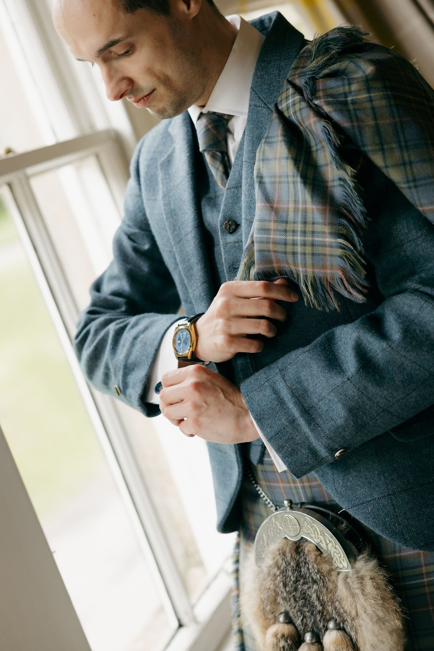 Groom in blue tweed suit and tartan kilt adjusting cufflinks by a window on his wedding day.