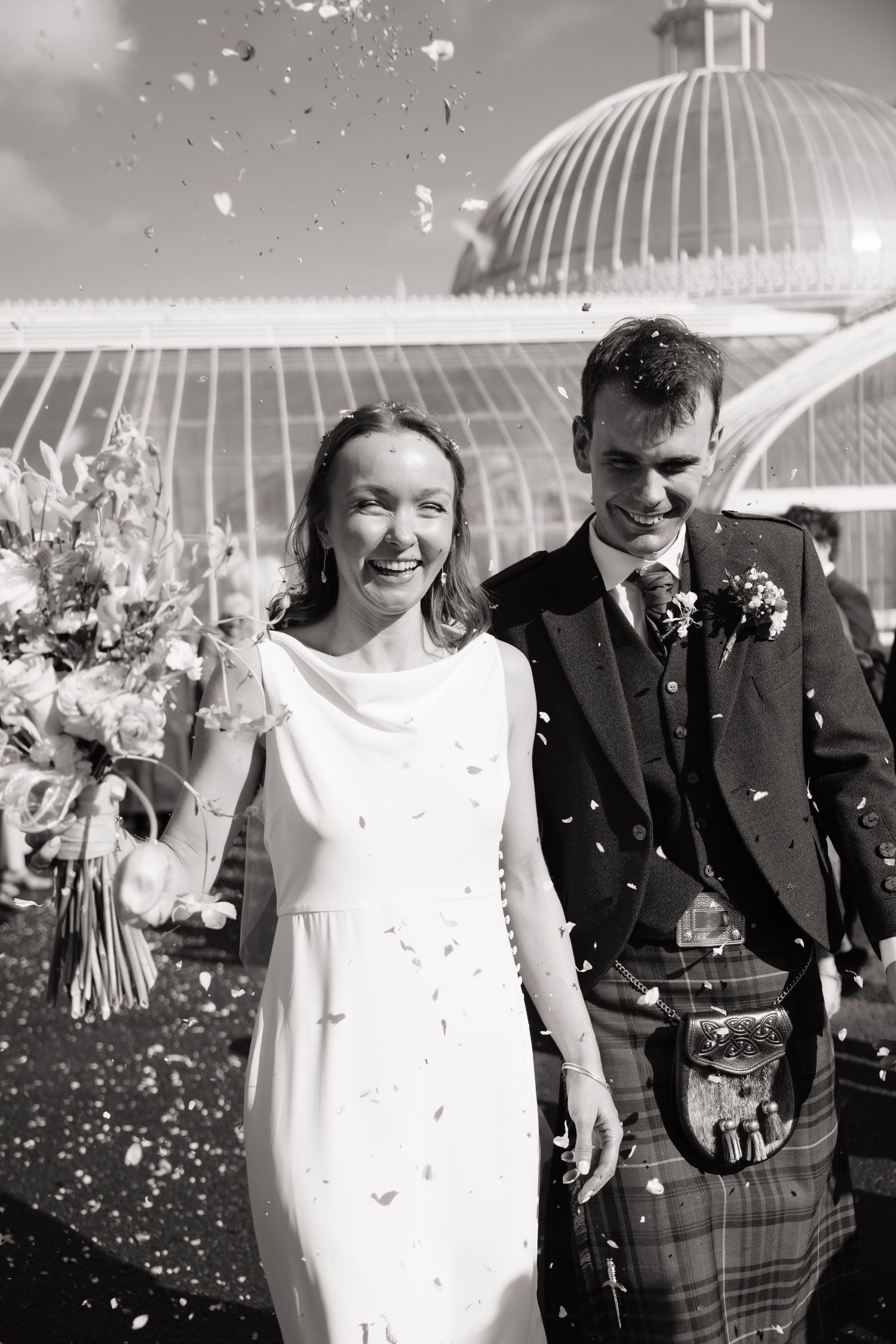 Black and white photo of smiling bride in white dress and groom in tartan kilt celebrating with confetti outside a greenhouse.