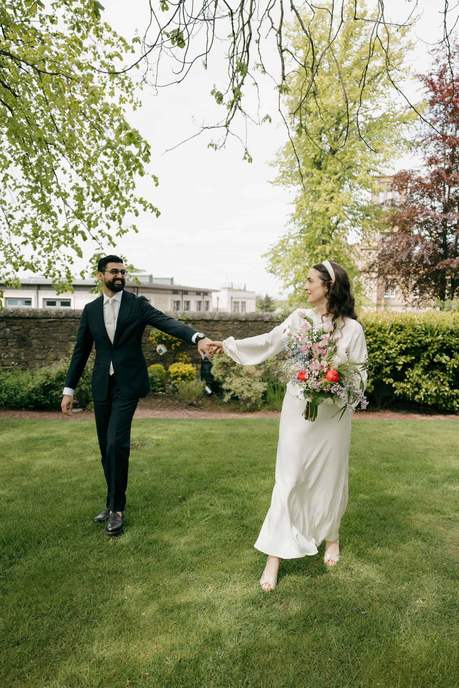 Bride and groom holding hands walking on green lawn in garden, bride in white dress holding colorful bouquet.