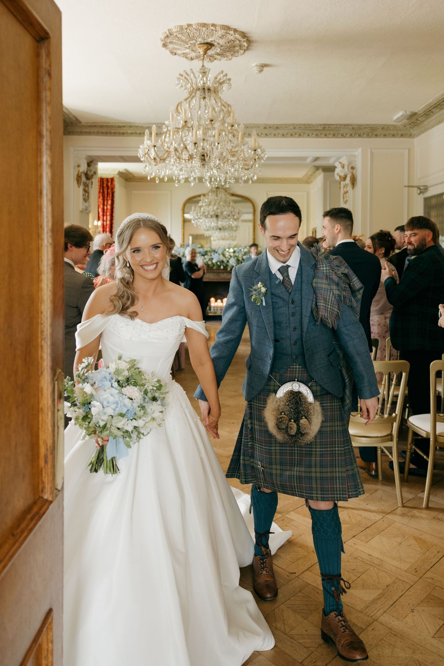 Smiling newlyweds walk hand-in-hand after ceremony, groom in Scottish tartan kilt under chandelier.