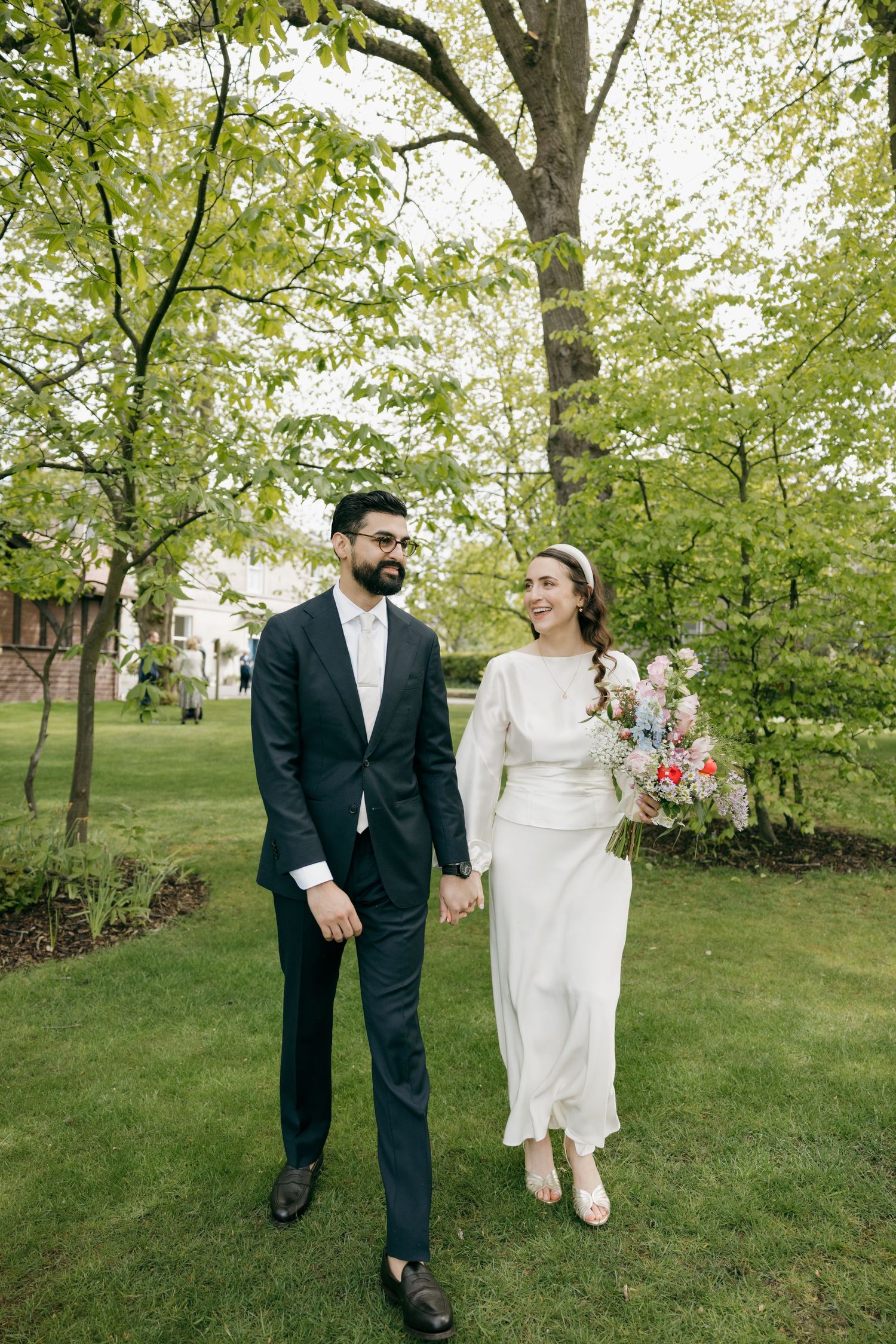 A bride and groom hold hands walking through a lush green park, she in a white gown, he in a navy suit.