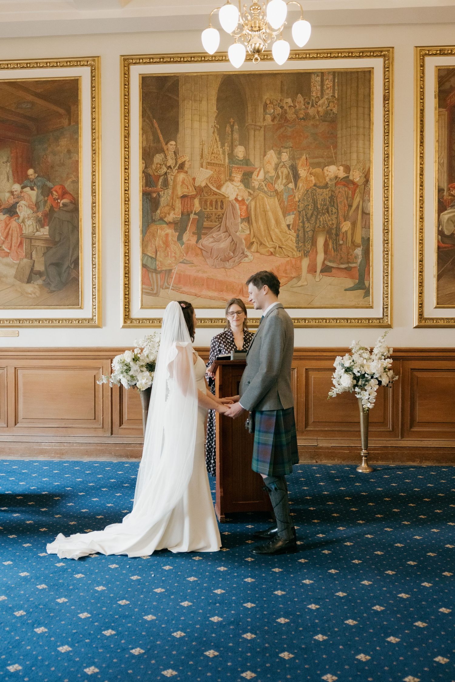Bride and groom exchange vows in an elegant Scottish hall with historic paintings and blue carpet.
