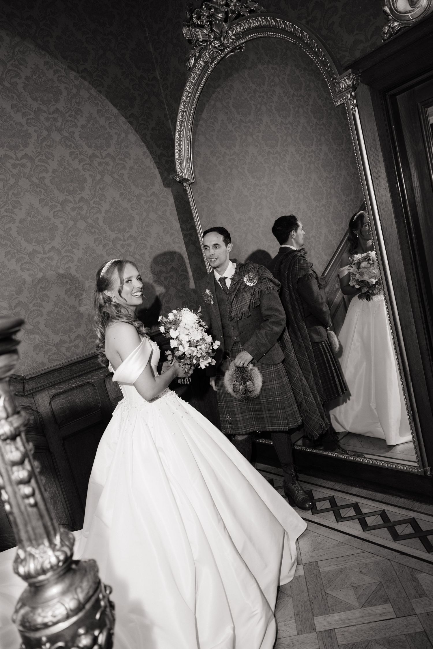 Black and white photo of bride and groom in kilt reflected in ornate gold mirror after ceremony.