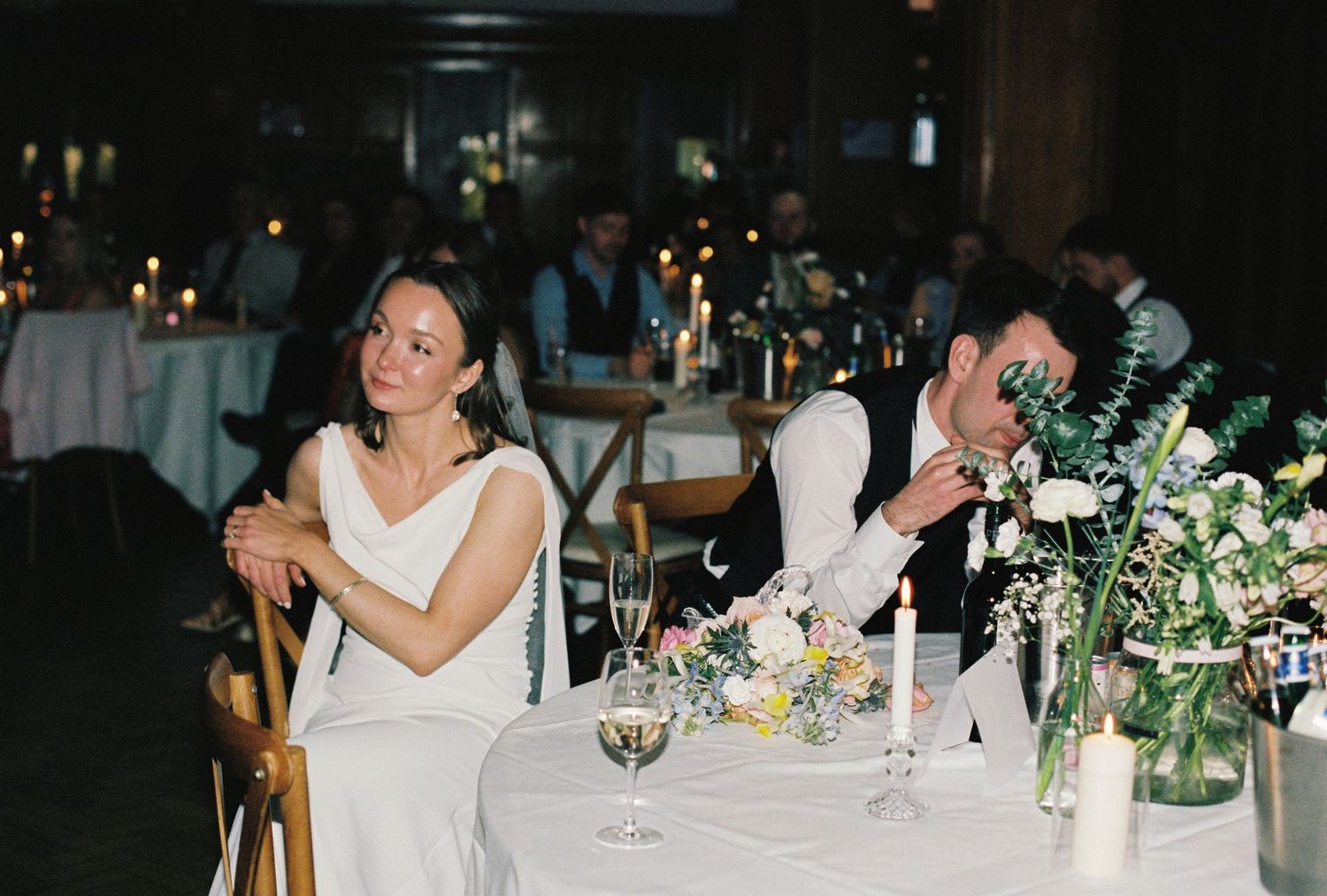 Bride and groom seated at wedding reception table with candles and floral centerpieces in dimly lit venue.