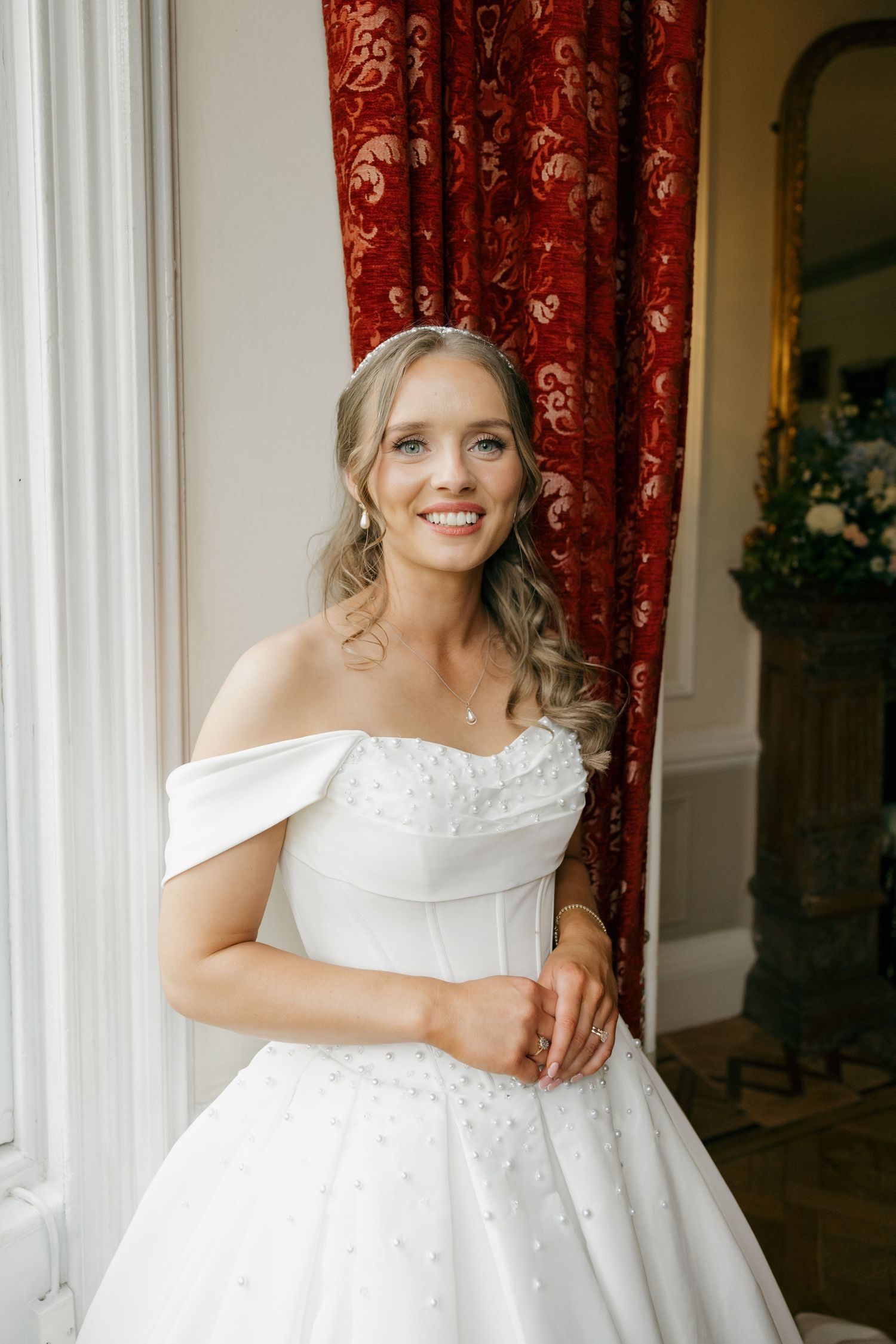 Smiling bride in an off-shoulder white ballgown standing by a window with red curtains.