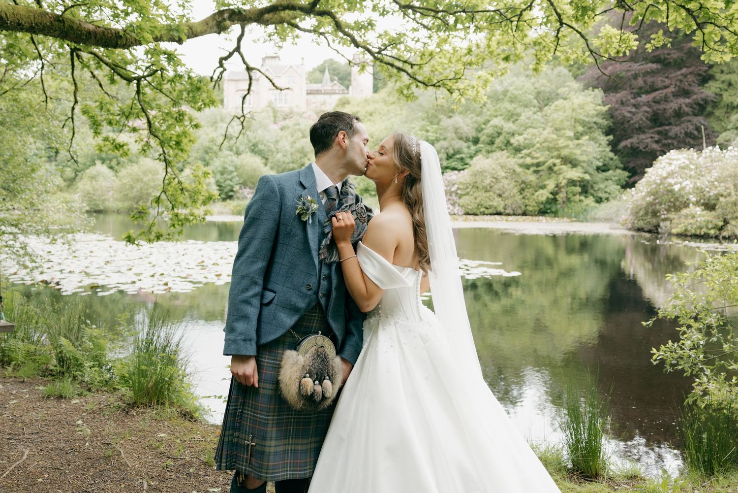 Newlyweds share a romantic kiss by a serene loch, groom wearing traditional Scottish tartan kilt.