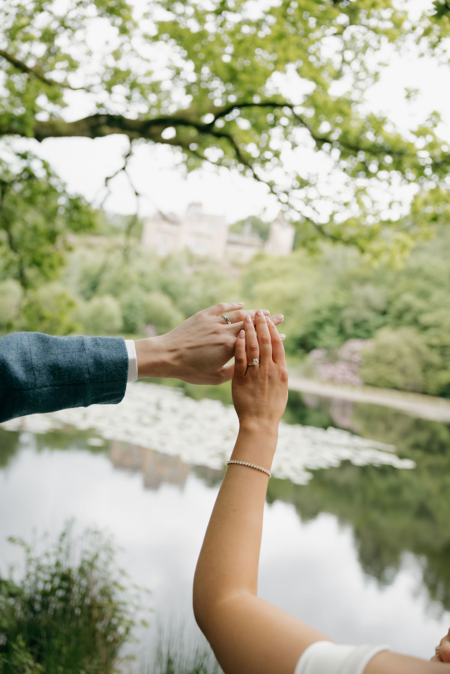 Bride and groom hold hands over a serene lily pond surrounded by lush green trees at their wedding.