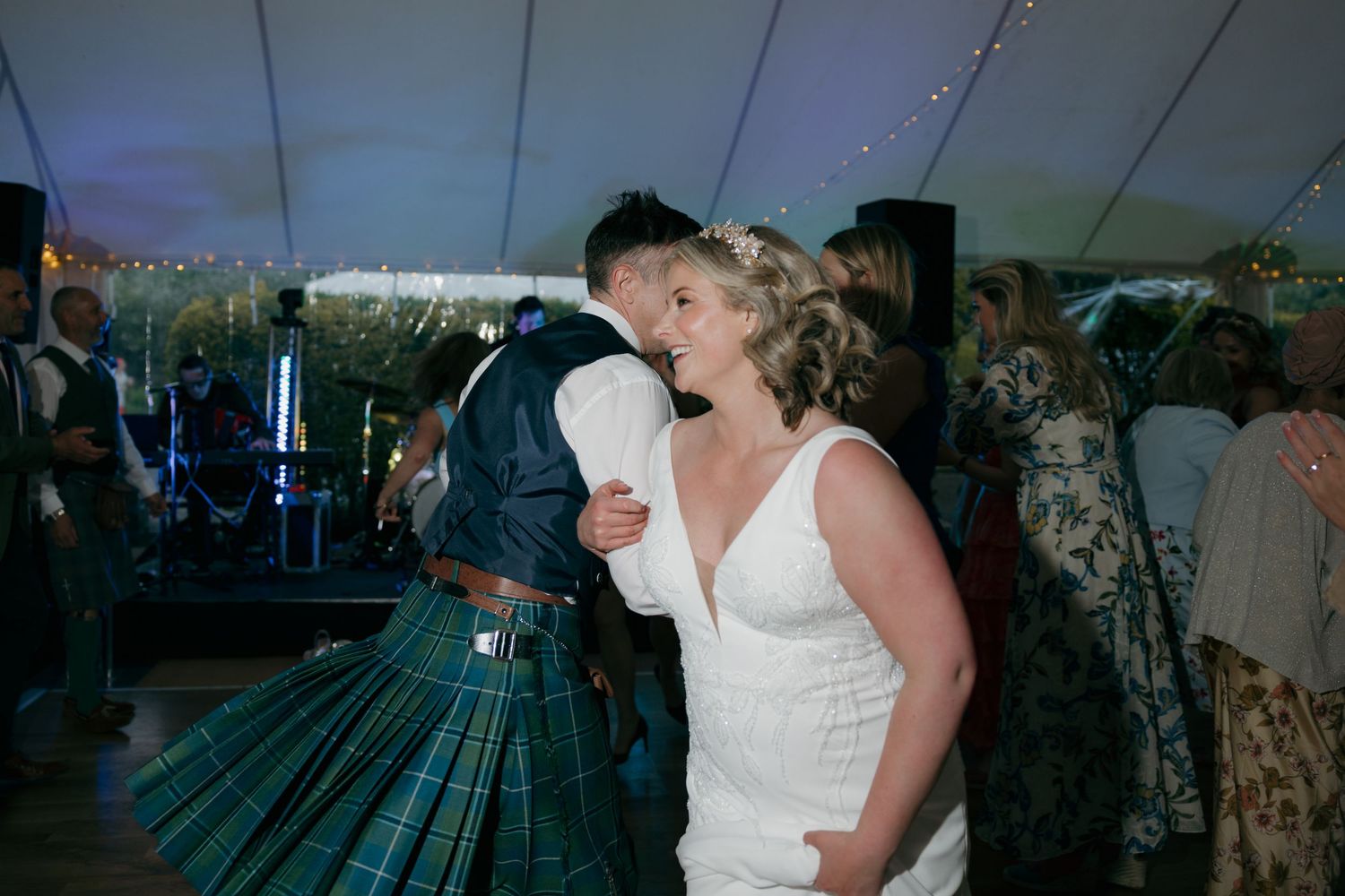 Bride in white lace dress dancing with groom in traditional Scottish tartan kilt at wedding reception.