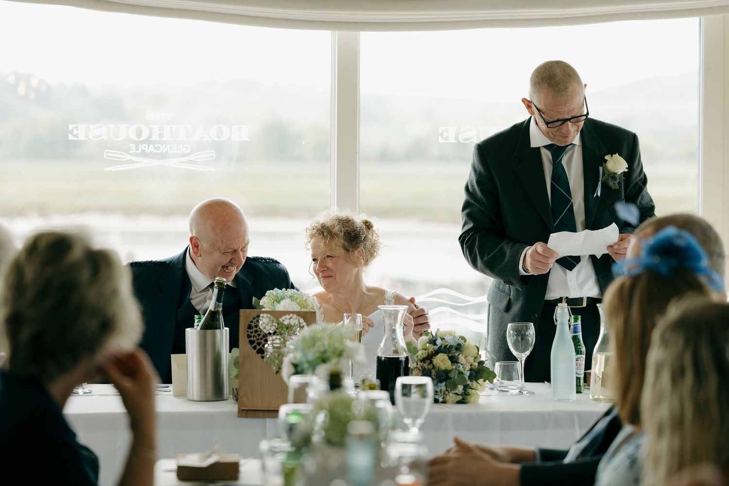 Groom reads wedding speech at reception table while bride and guests smile and laugh in bright venue.