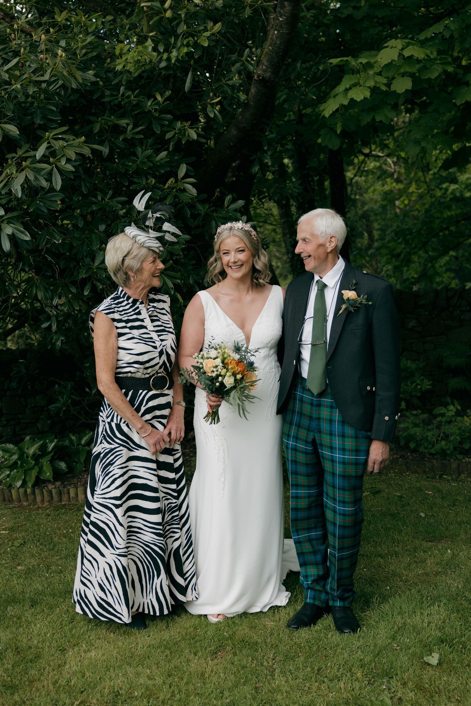 Bride in white gown poses with parents, mother in zebra print dress and father in blue tartan kilt, outdoors.