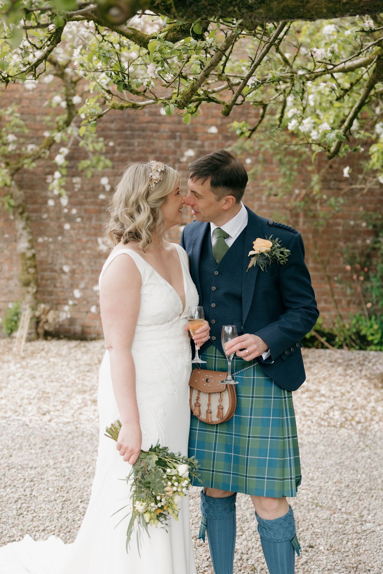 Bride and groom in Scottish kilt share a romantic moment holding champagne flutes and bouquet in a walled garden.