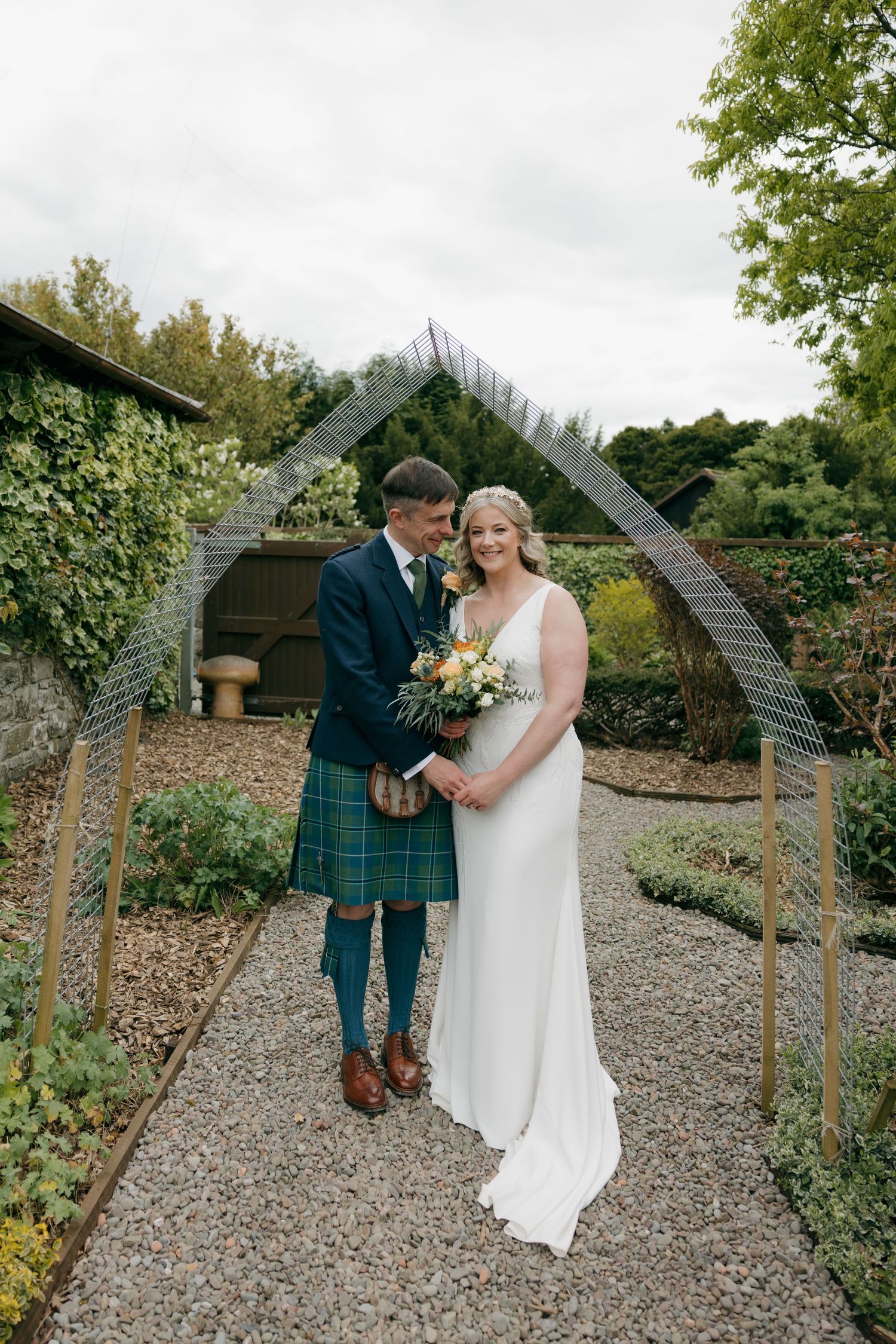 Bride and groom pose under wire arch in garden, groom wearing blue tartan kilt, bride holding colorful bouquet.